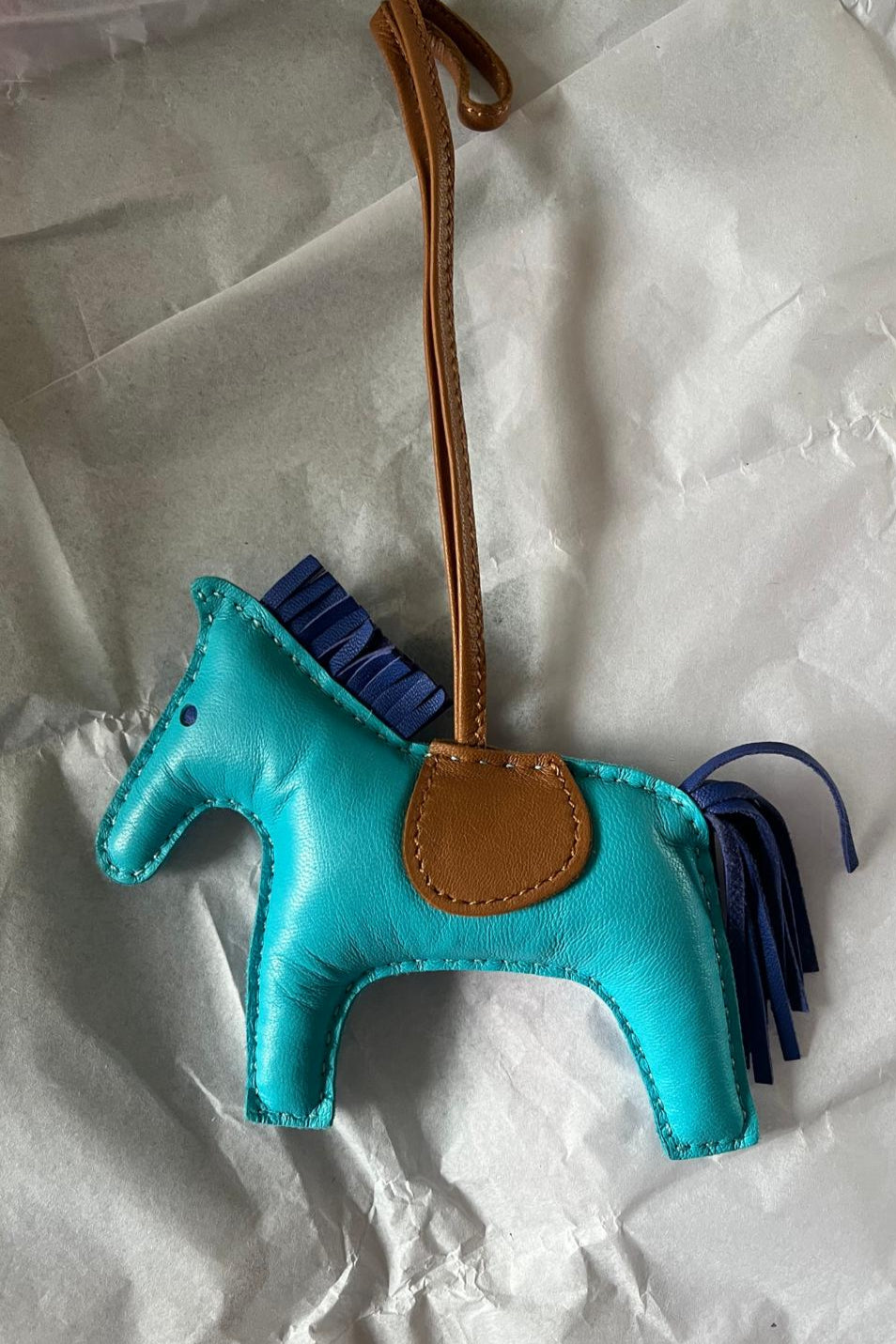 Turquoise horse-shaped bag charm with brown leather strap on crumpled white paper
