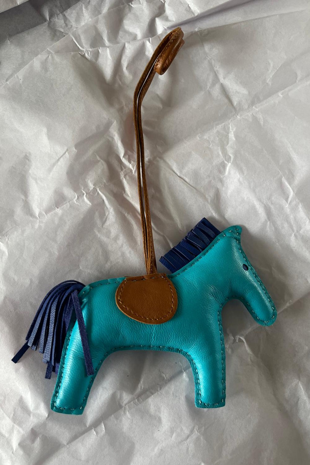 Turquoise horse-shaped bag charm with brown leather strap on crumpled white paper