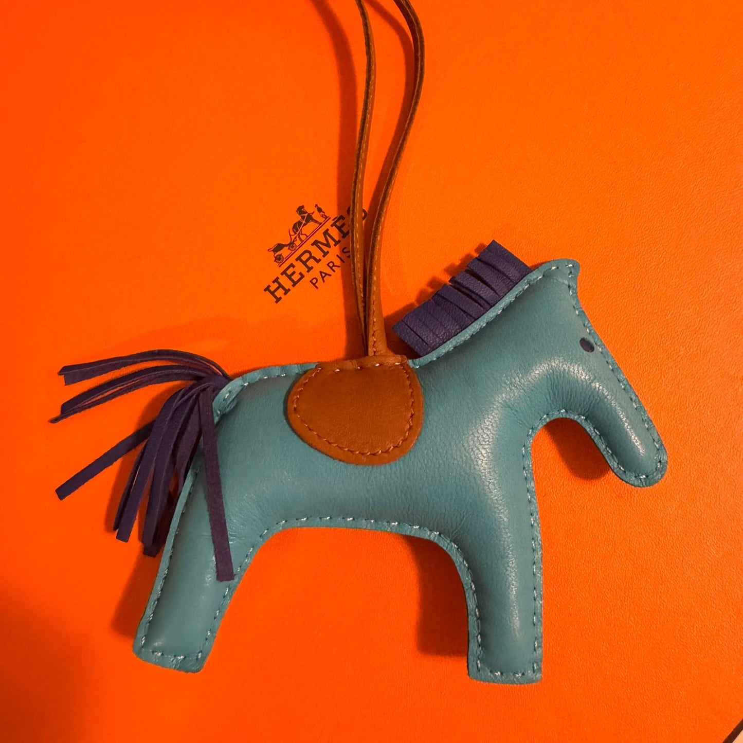 Blue horse-shaped bag charm with brown leather patch on an orange background