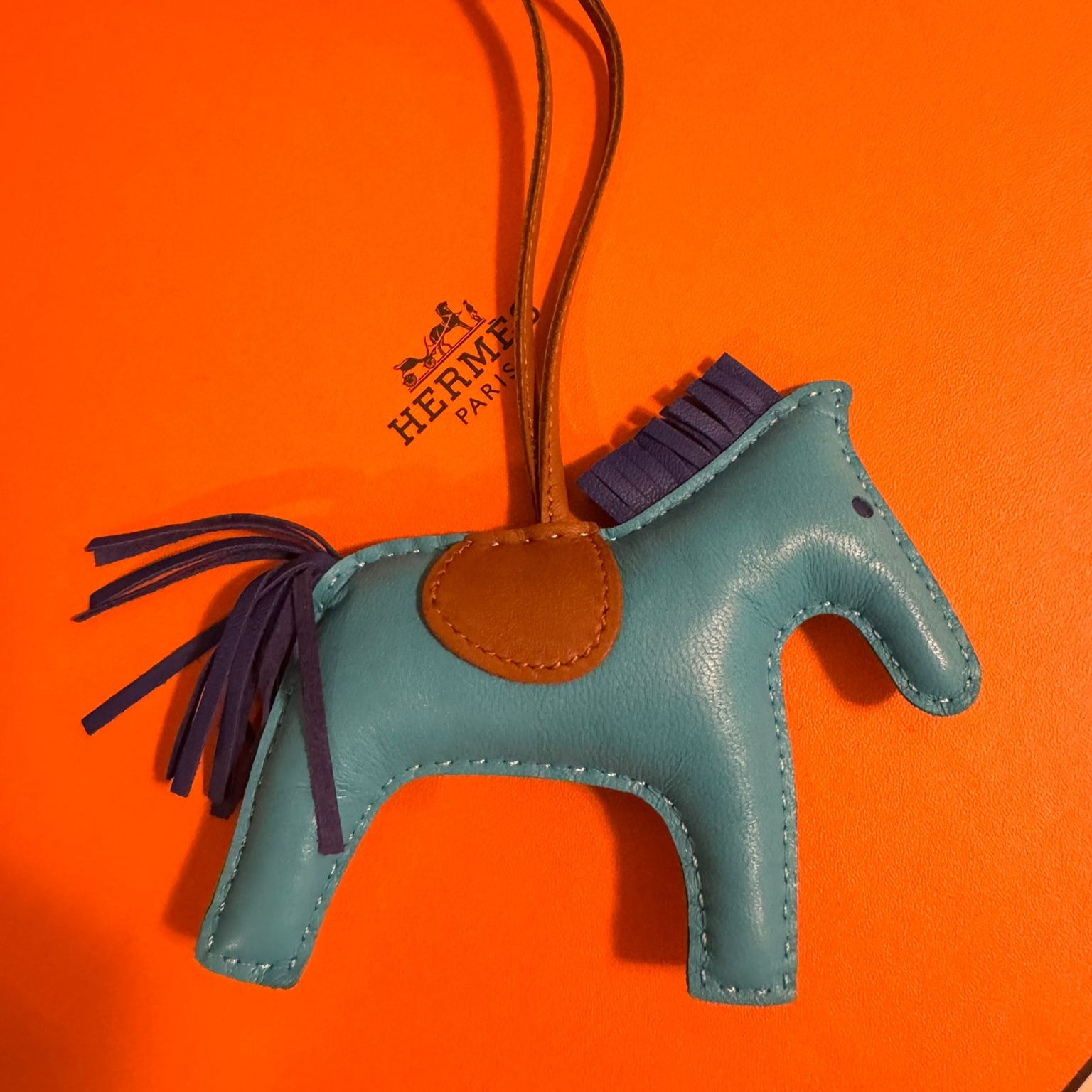 Blue horse-shaped bag charm with brown leather patch on an orange background